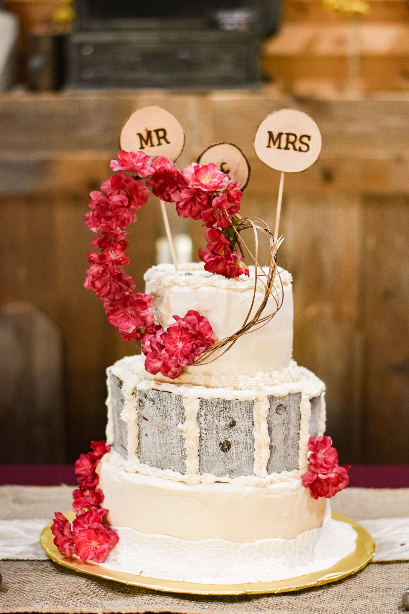 Three-tier wedding cake decorated with red flowers and Mr & Mrs toppers on wooden table.