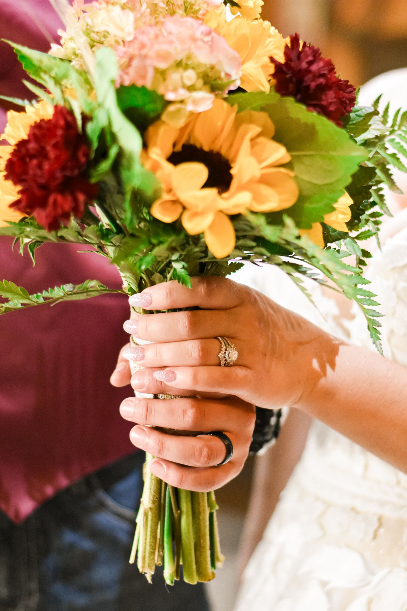 Wedding bouquet with sunflowers and burgundy flowers held by hands showing engagement ring.