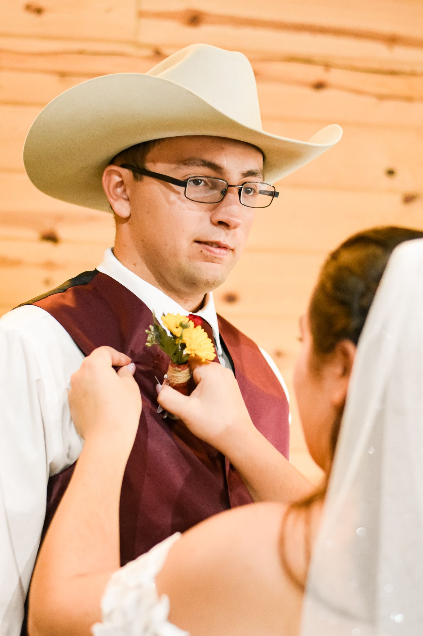Wedding moment with groom in cowboy hat and burgundy vest receiving boutonniere.