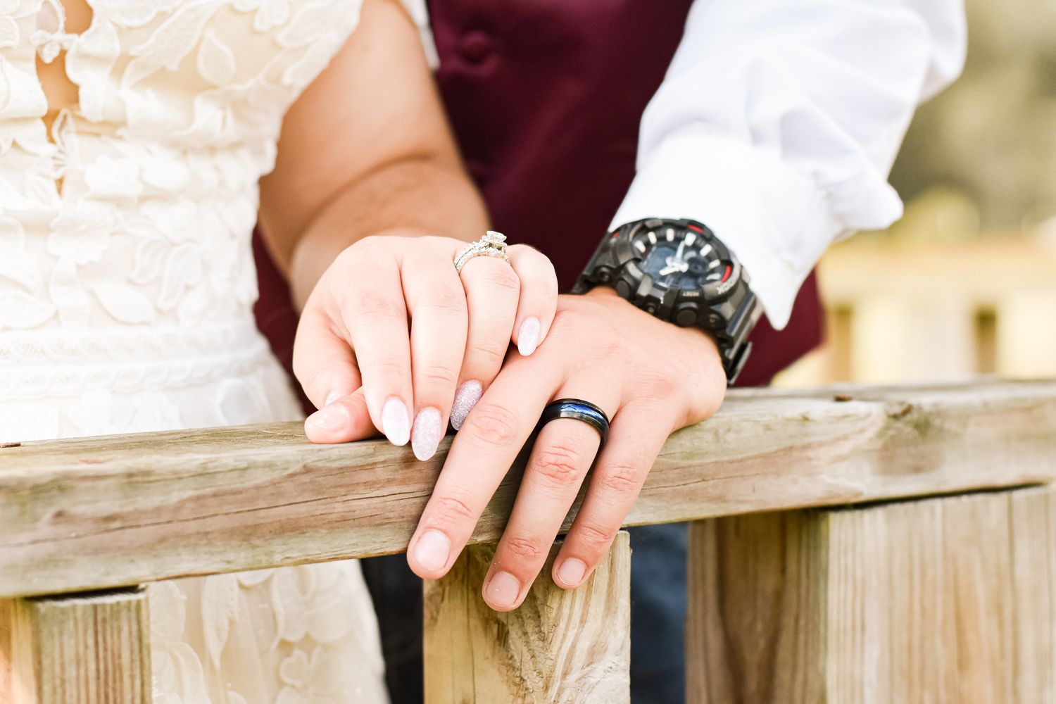 Close-up of newlyweds' hands resting on wooden railing showing wedding rings and watch details.
