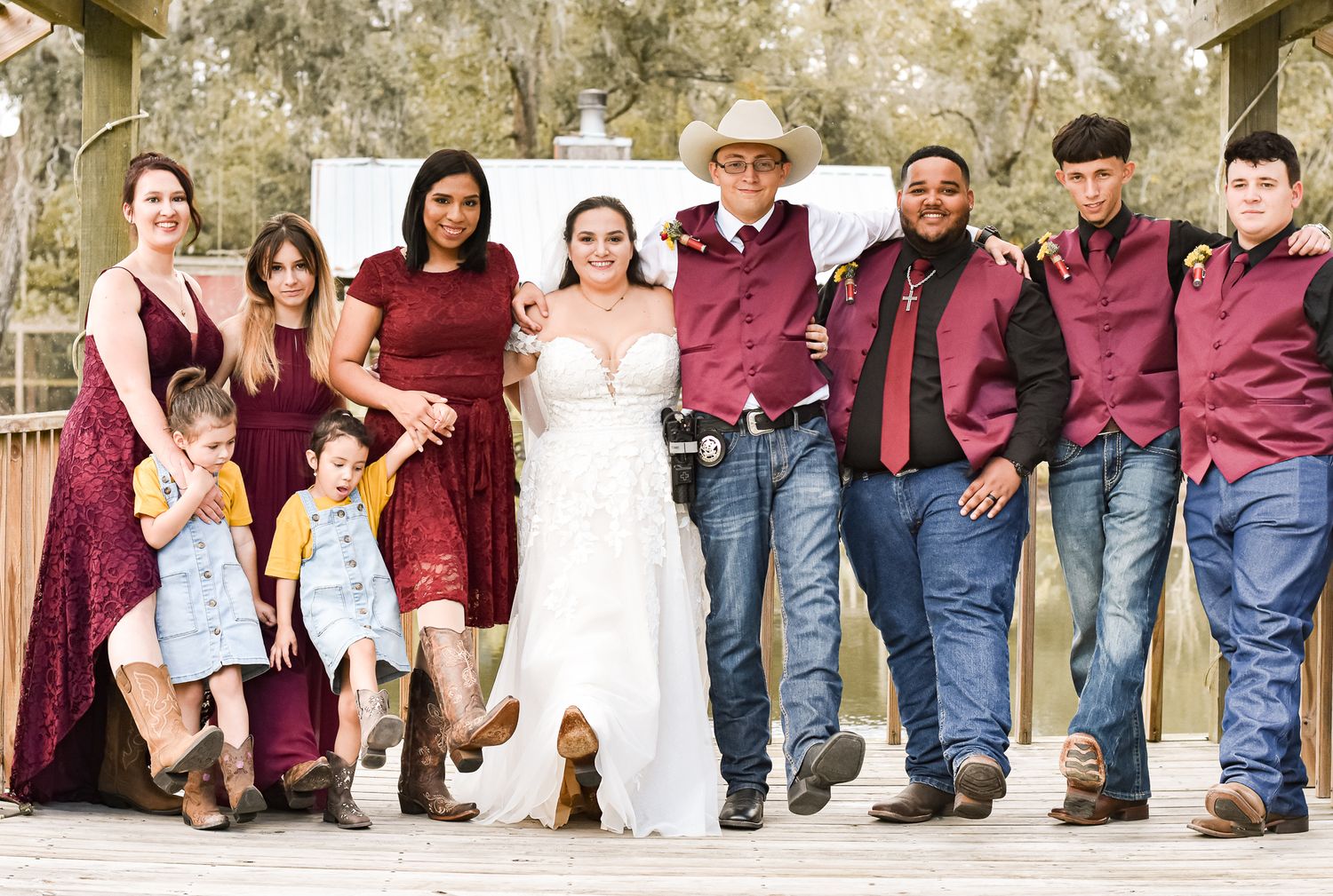 Wedding party in burgundy and denim attire poses on wooden deck.
