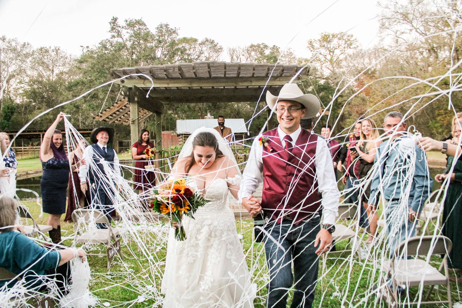 Wedding guests celebrate by throwing streamers as couple exits outdoor ceremony under wooden pergola.