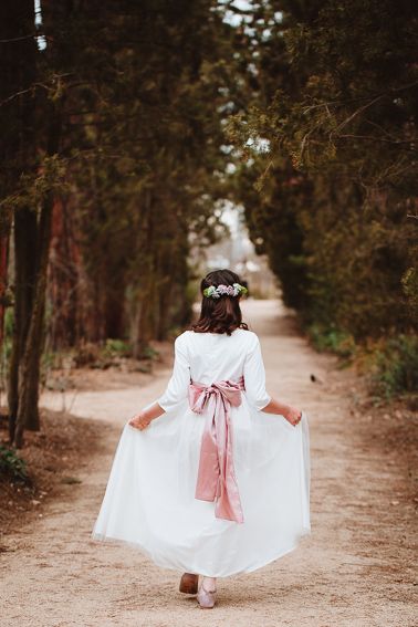 Back view of person in white dress with pink bow walking down a tree-lined path.
