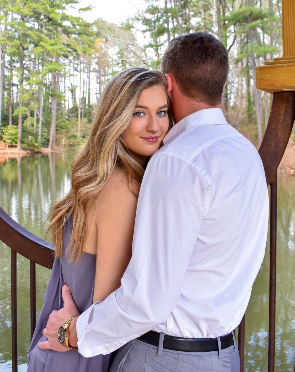 Romantic couple embracing on wooden deck overlooking pond with pine trees in background.