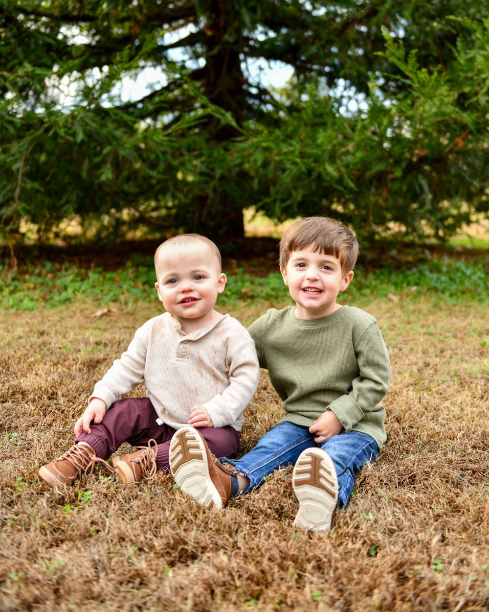 Two young boys sit together on grass wearing matching neutral-colored sweaters.