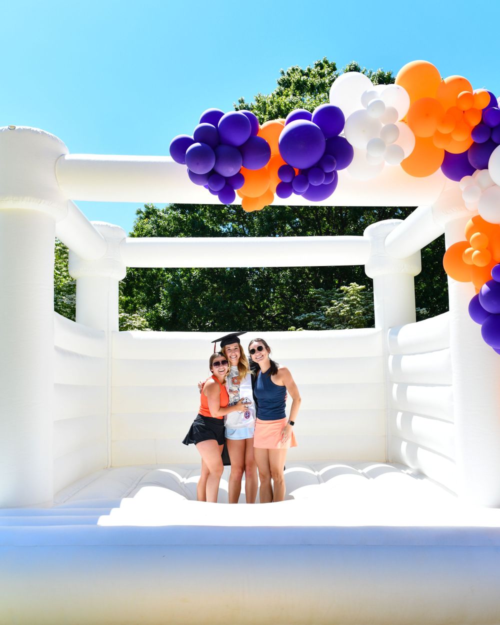 Three friends pose inside white bounce house decorated with purple and orange balloons.