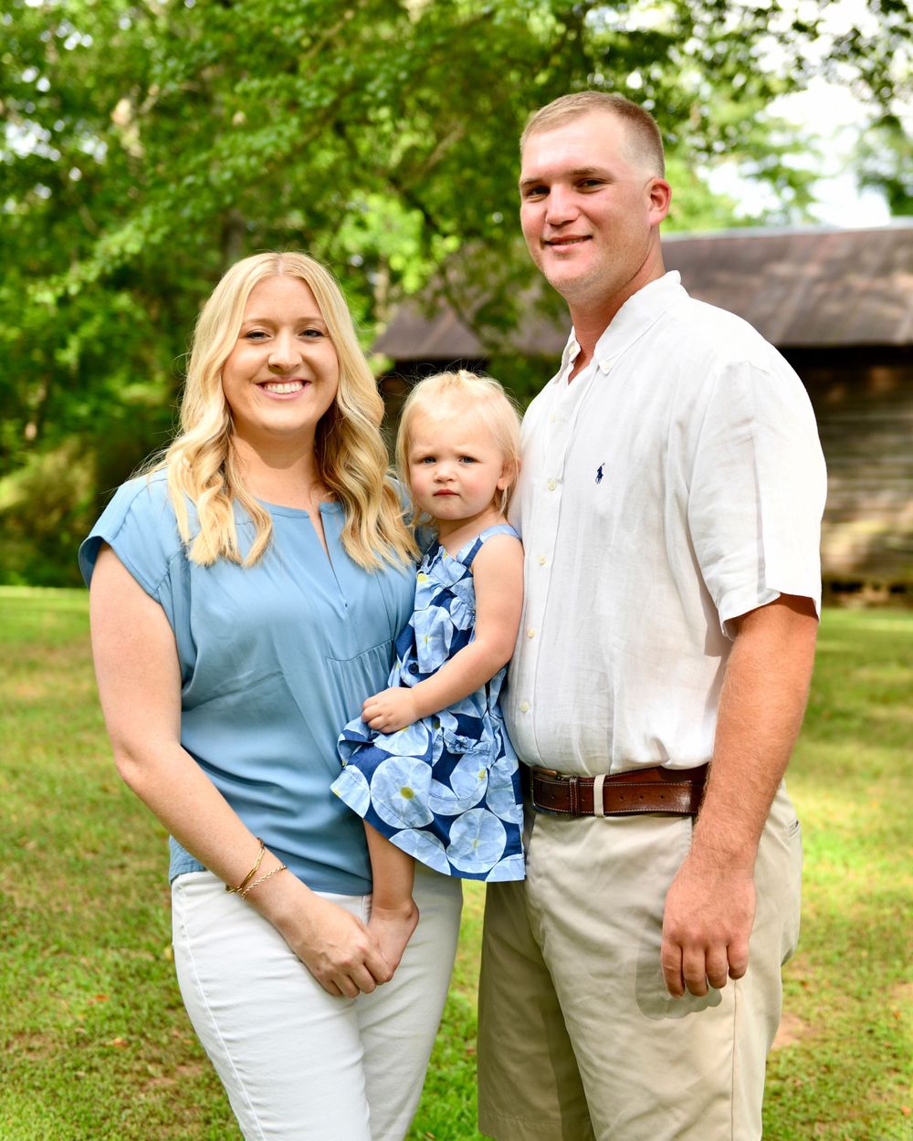 A family portrait taken outdoors with greenery and a rustic building in the background.