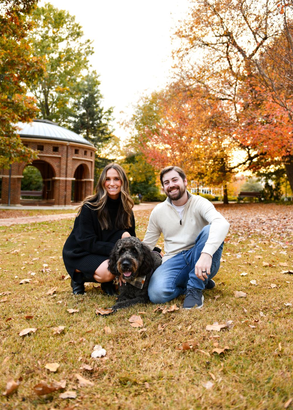 Couple and their black dog pose for a photo in front of brick building during fall.