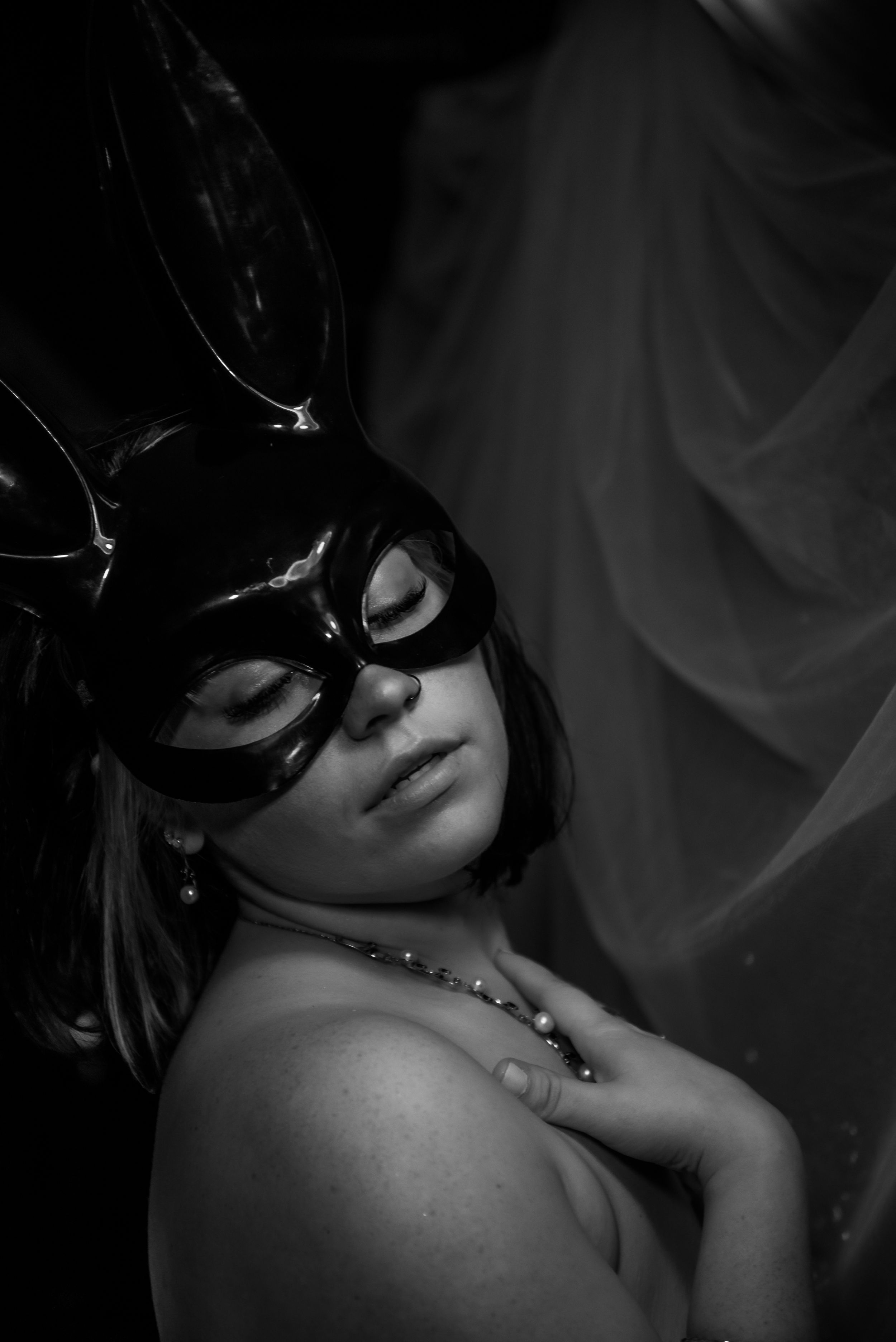 boudoir image black and white image portrait of woman clutching necklace wearing a black plastic half faced bunny mask white drapes behind