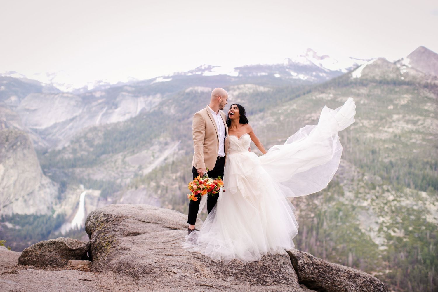 Groom in khaki suit jacket and bride in flowing dress standing on Yosemite National Park cliff