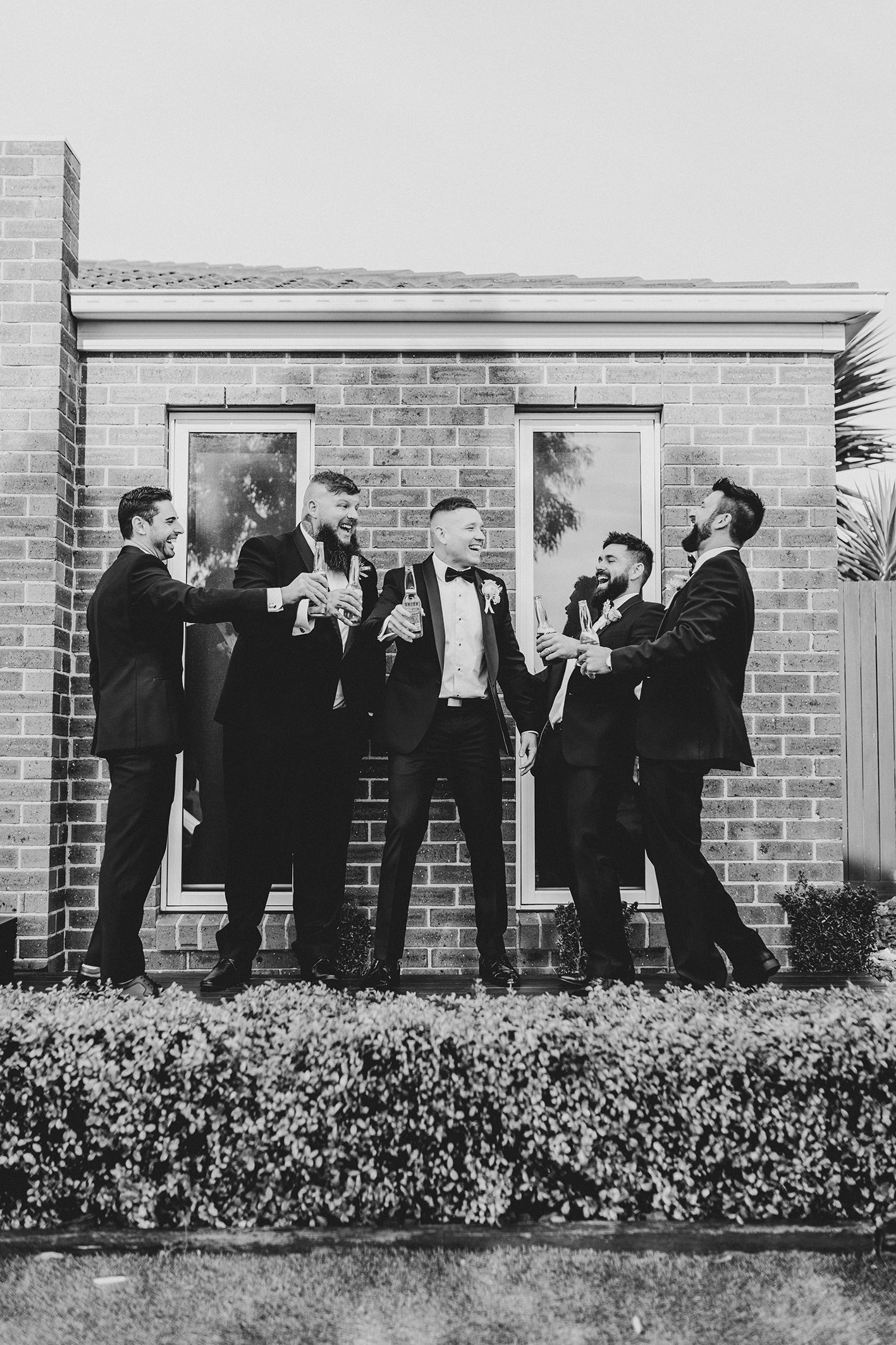 Groomsmen in black tuxedos pose playfully against a brick building exterior.