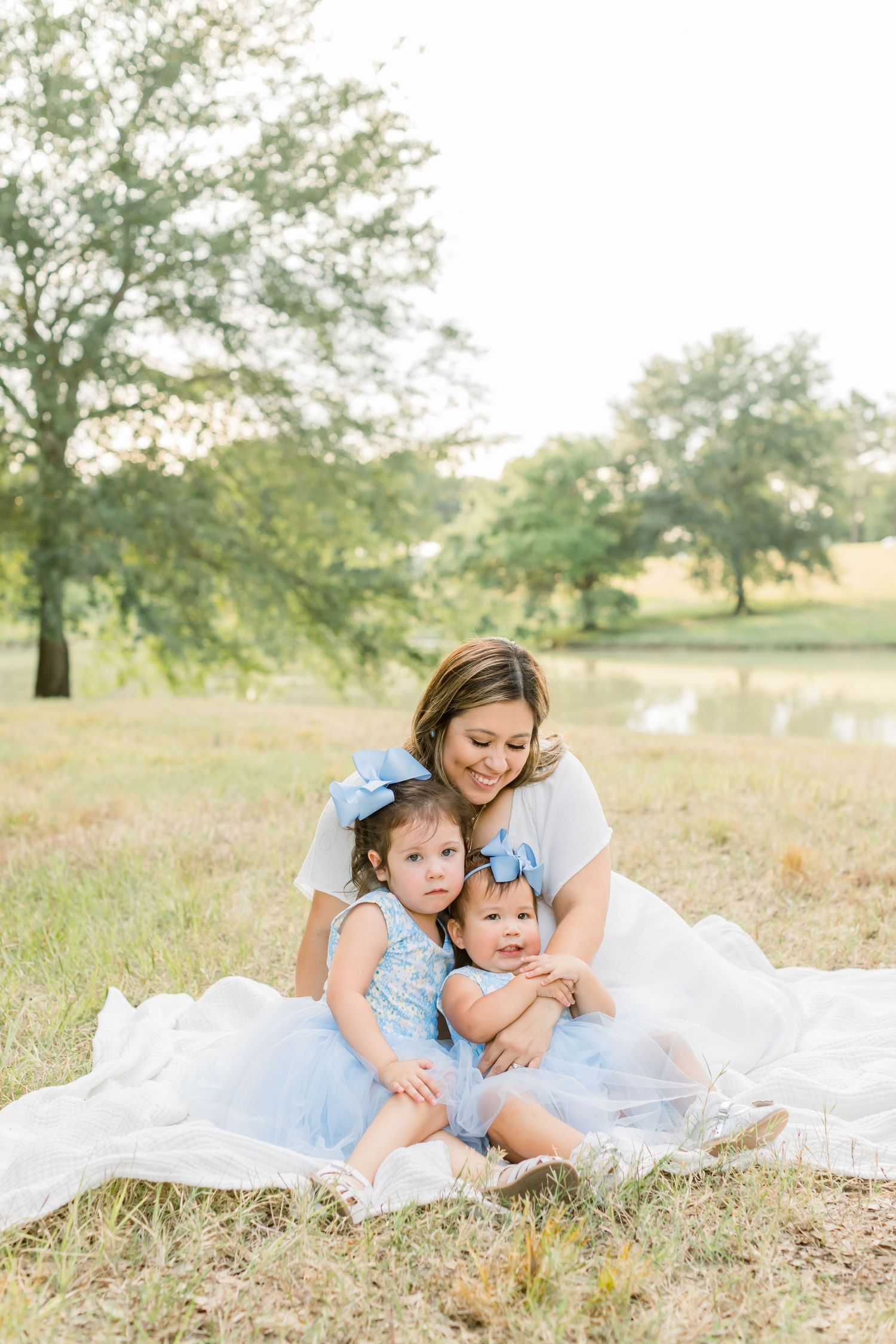 Mother cuddling with children in blue dresses on white blanket outdoors.