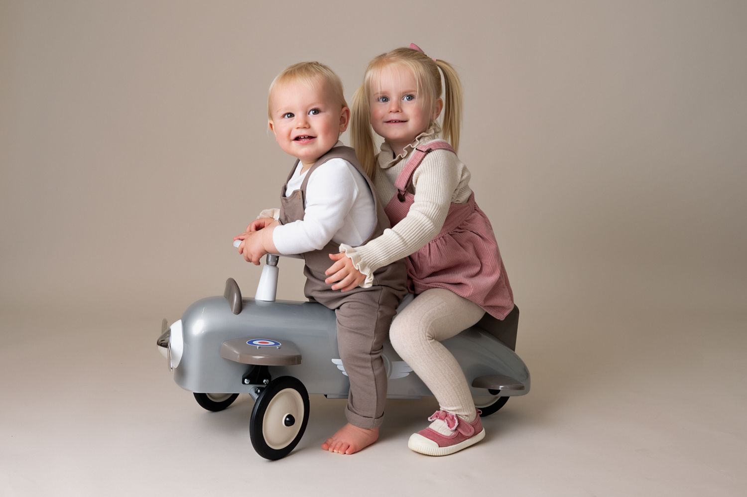 Two children riding a vintage toy airplane on neutral studio background.