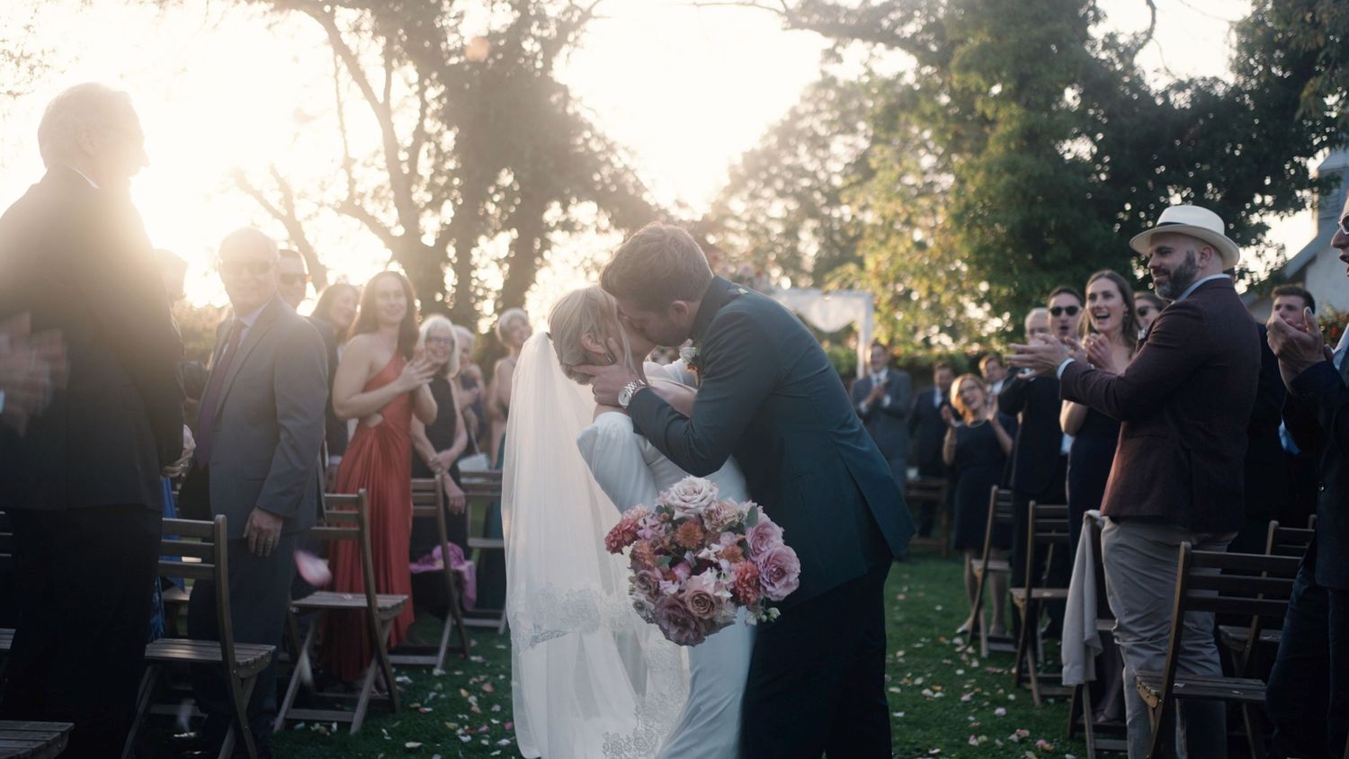 Outdoor wedding ceremony with guests watching couple exchange vows under sunlit trees.