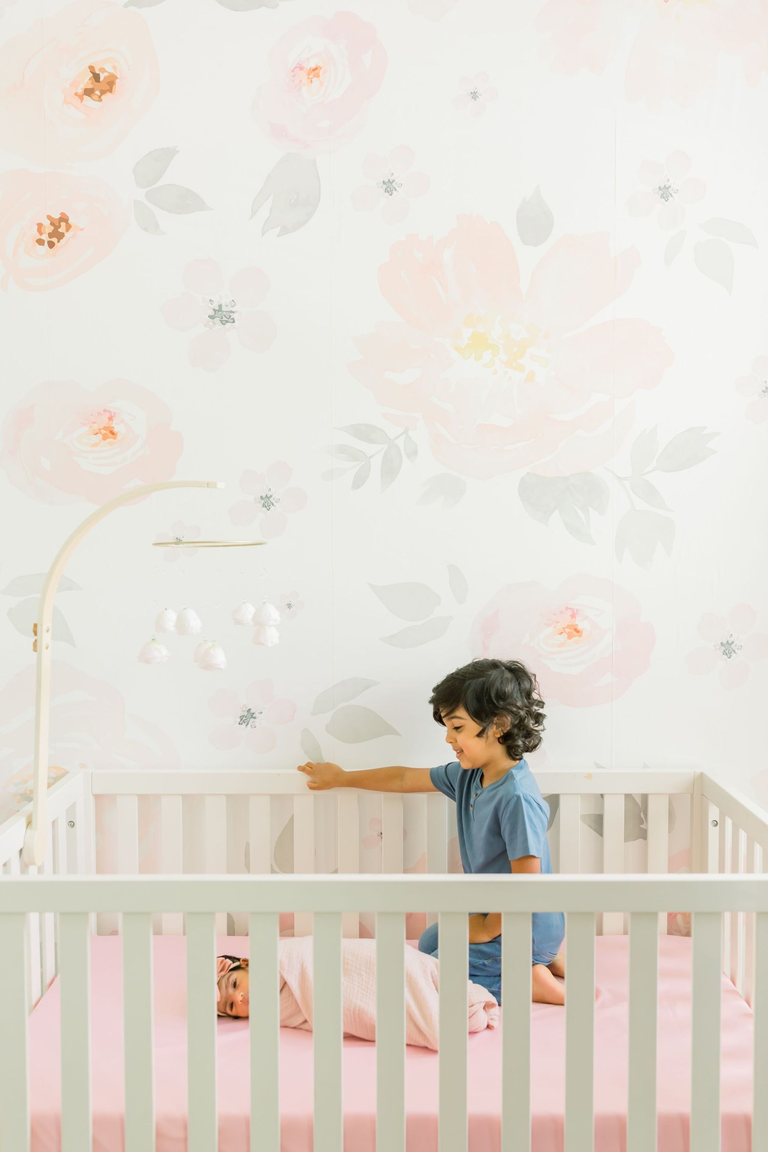 Child looking over white crib railing against floral nursery wallpaper.