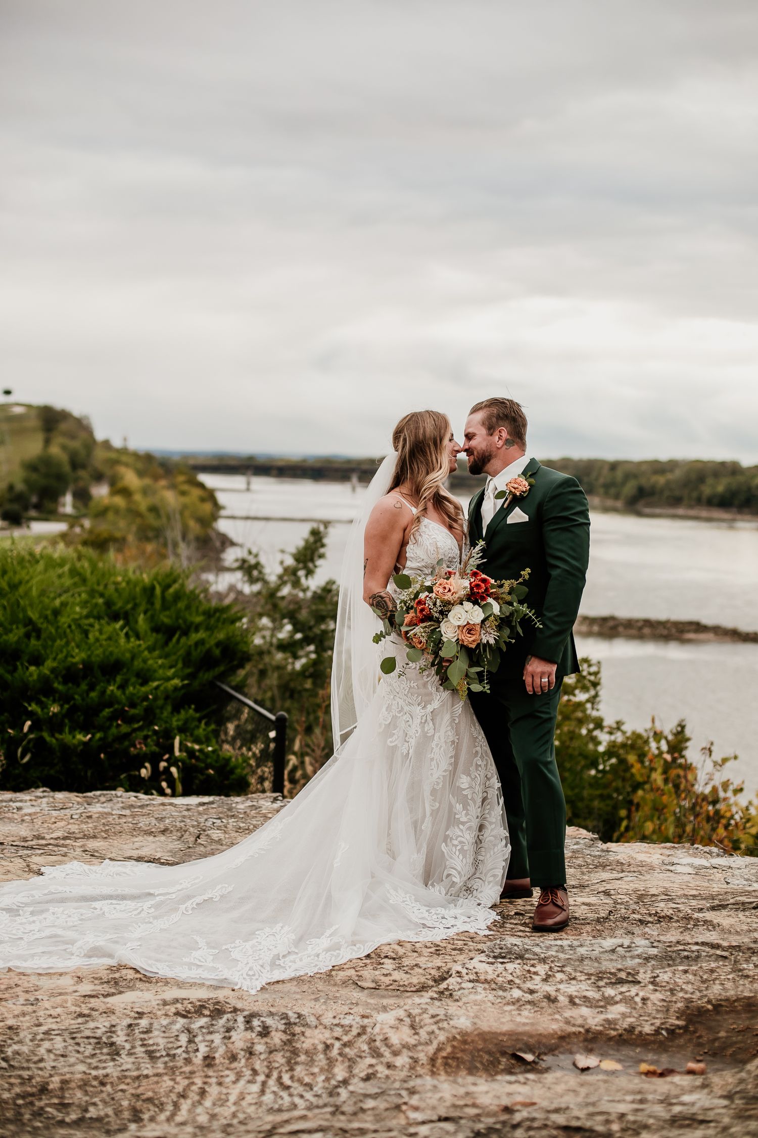 Newlyweds share an intimate moment on rocky terrain overlooking a scenic waterfront.