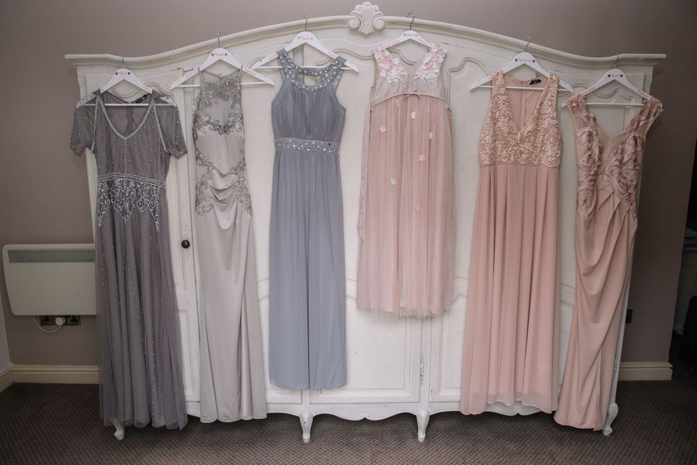 Collection of bridesmaid dresses in soft pink and grey tones hanging on ornate white wardrobe.
