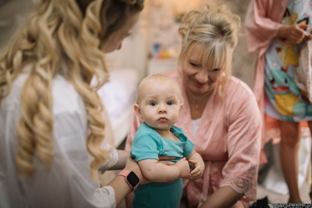 Baby in turquoise outfit being held during a gathering of people in soft pink clothing.