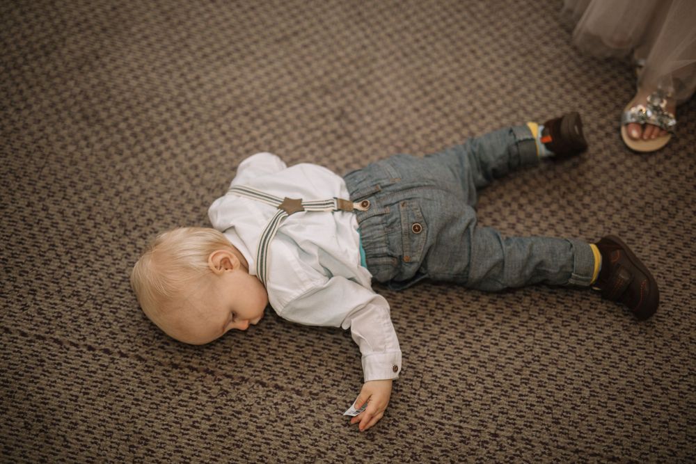A small child in white clothing and jeans lying on brown carpeted floor.