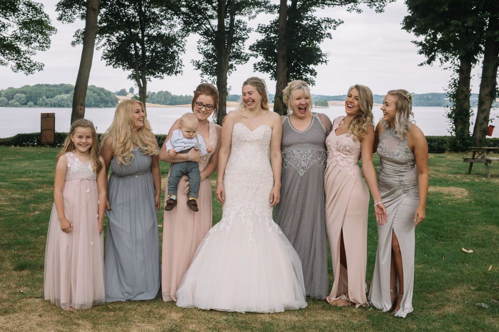 Wedding party in pastel dresses pose together on grassy lawn with lake and trees in background.