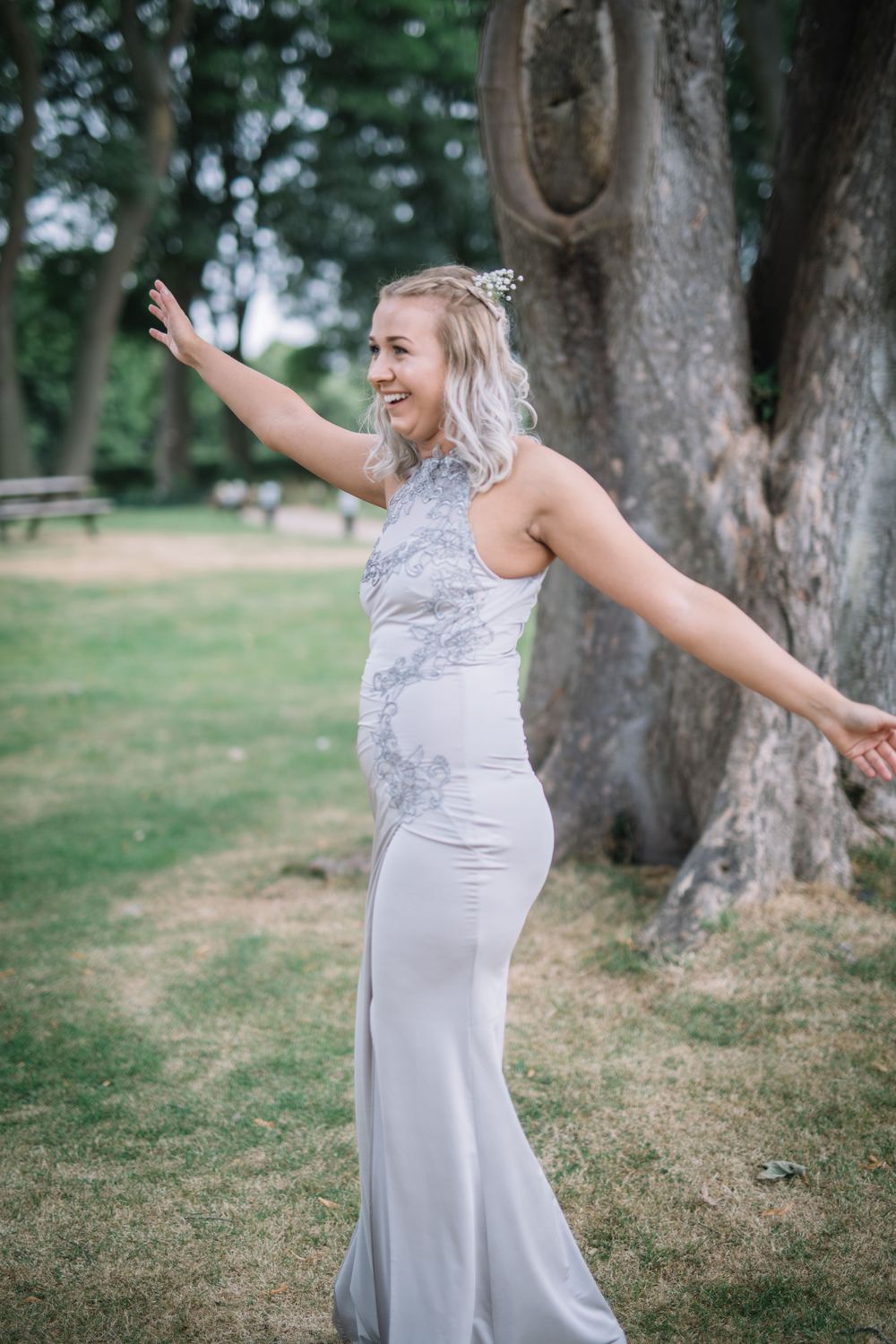 Woman in silver fitted dress poses with arms outstretched outdoors near large tree.