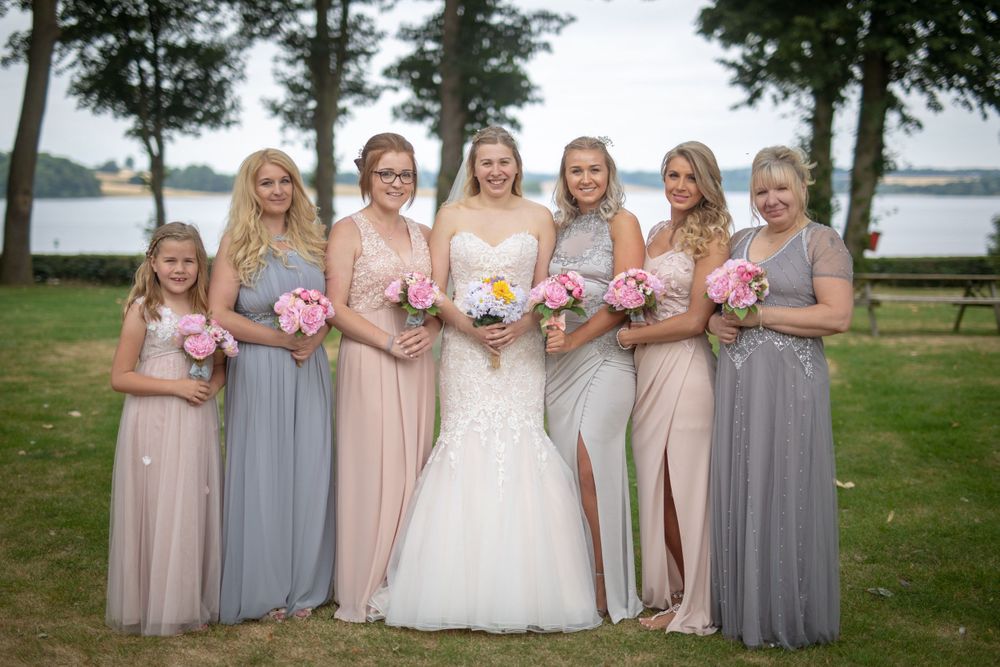 Bridal party wearing blush and grey gowns pose outdoors by a lake with coordinated bouquets.