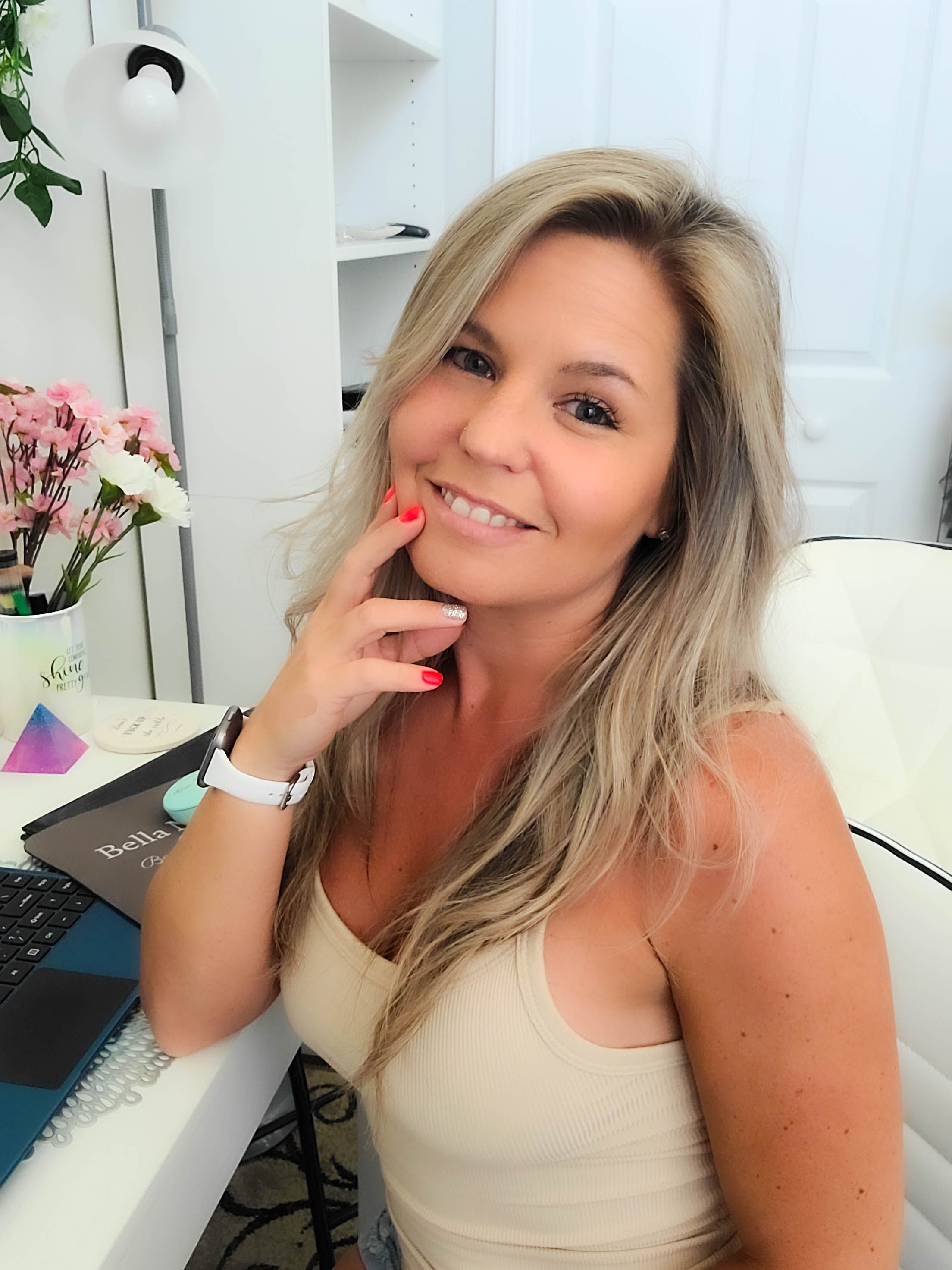 Woman in casual white tank top smiling at desk with flowers.