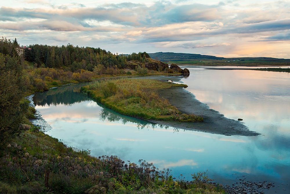 Serene lake landscape with green shoreline reflecting cloudy sky during golden hour.