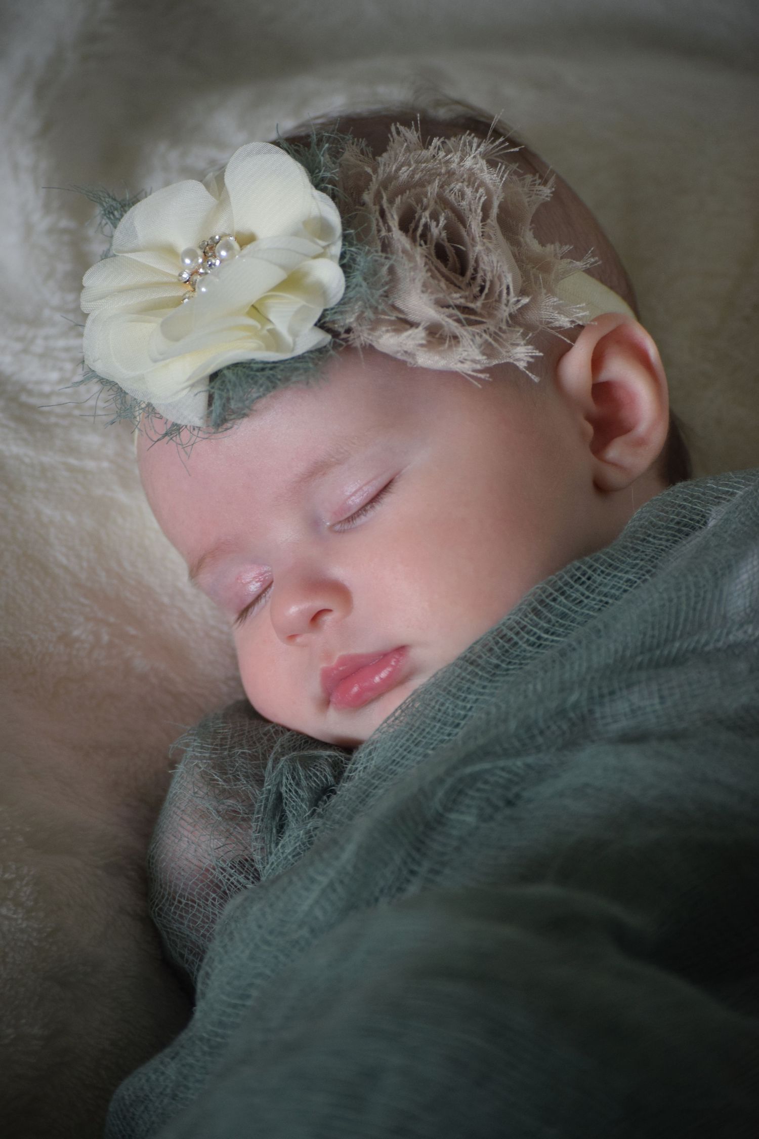 Peaceful sleeping newborn wearing sage swaddle and cream floral headband on soft white bedding.