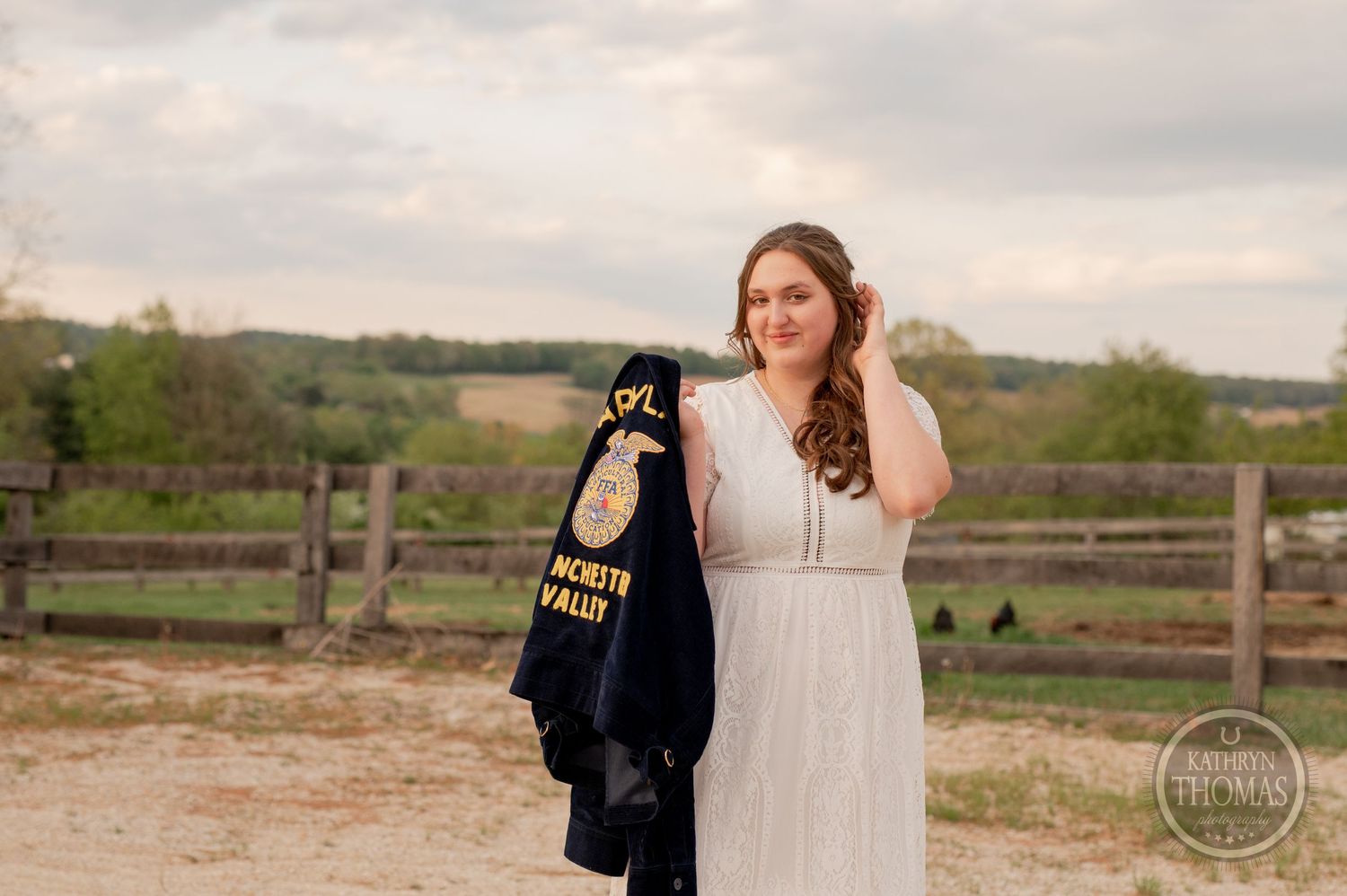 Someone in a white dress holds an FFA jacket while posing near a wooden fence at sunset.