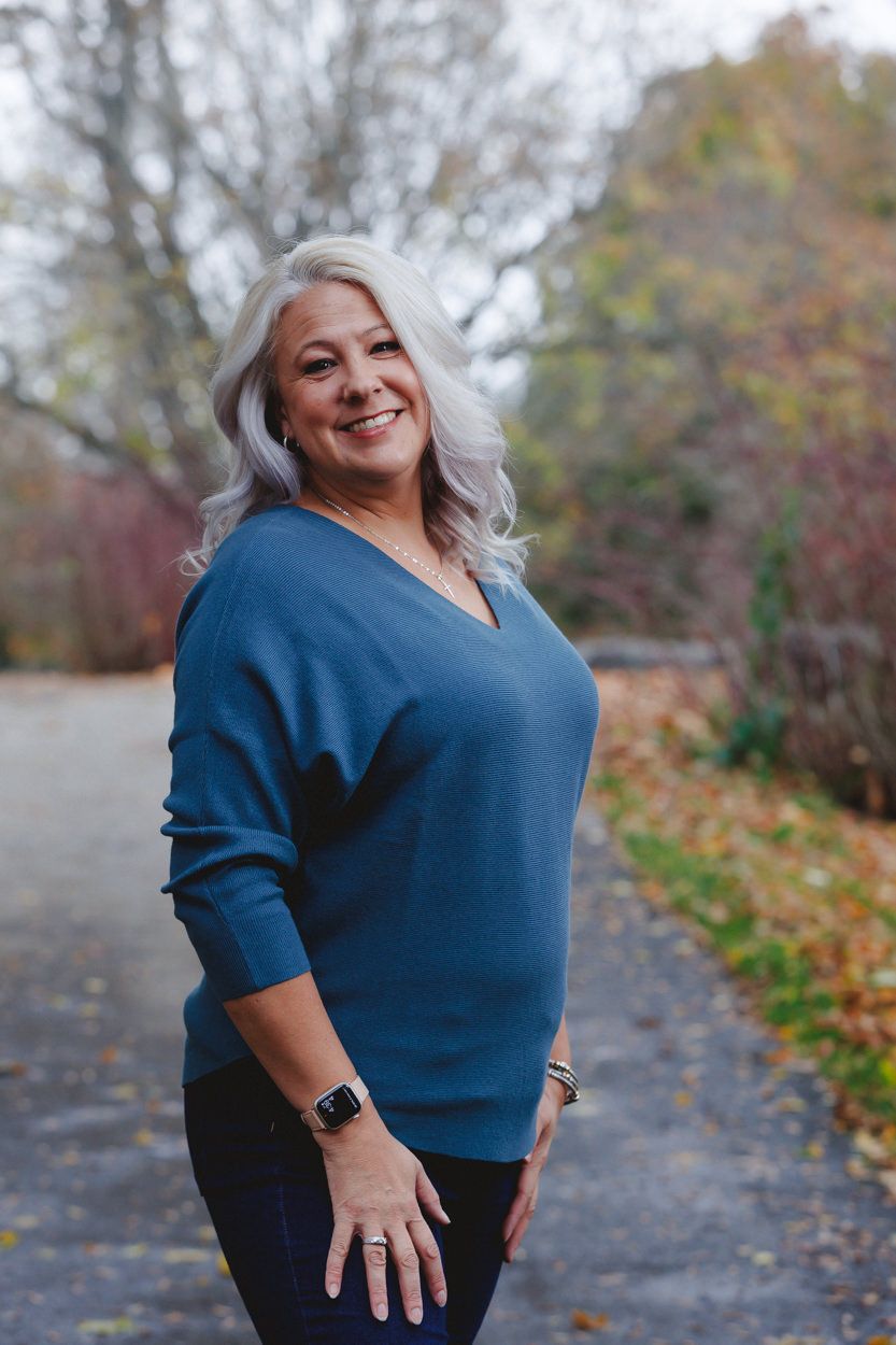 Woman in blue sweater poses outdoors on autumn path with fallen leaves.