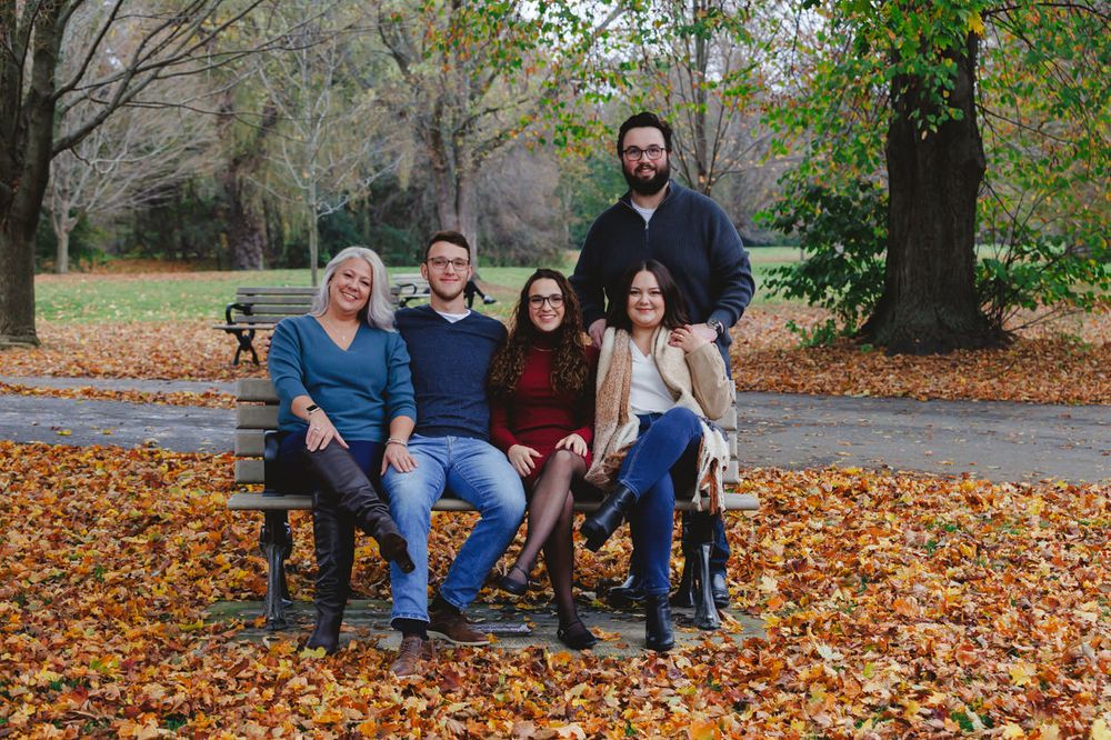 Family sitting together on wooden bench surrounded by fallen autumn leaves in park setting.