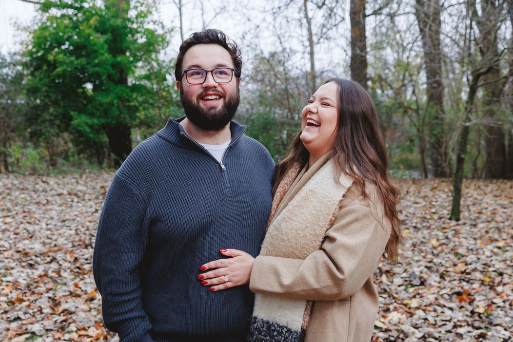 Couple laughing and embracing outdoors on an autumn day with fallen leaves in a wooded setting.