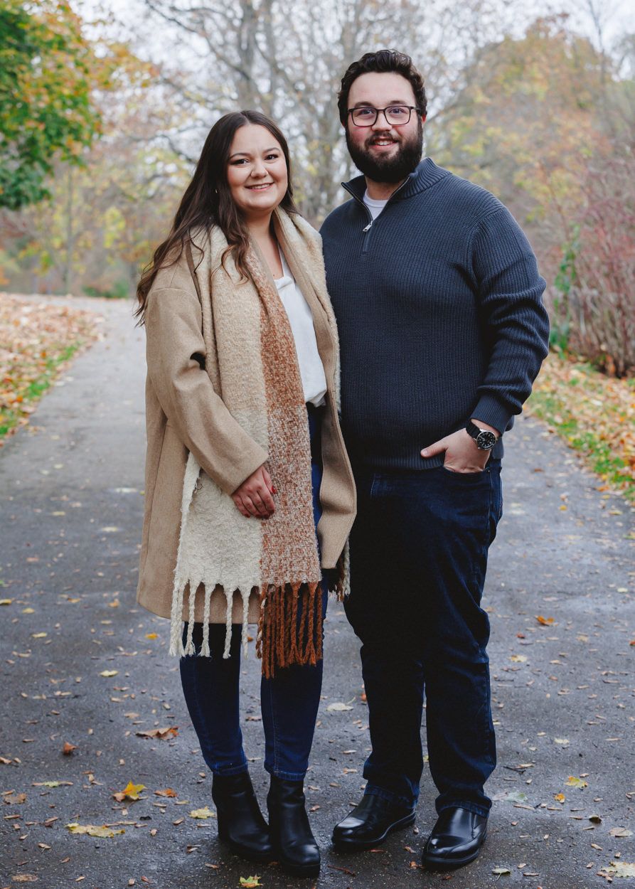 A couple stands together on a path surrounded by autumn foliage.
