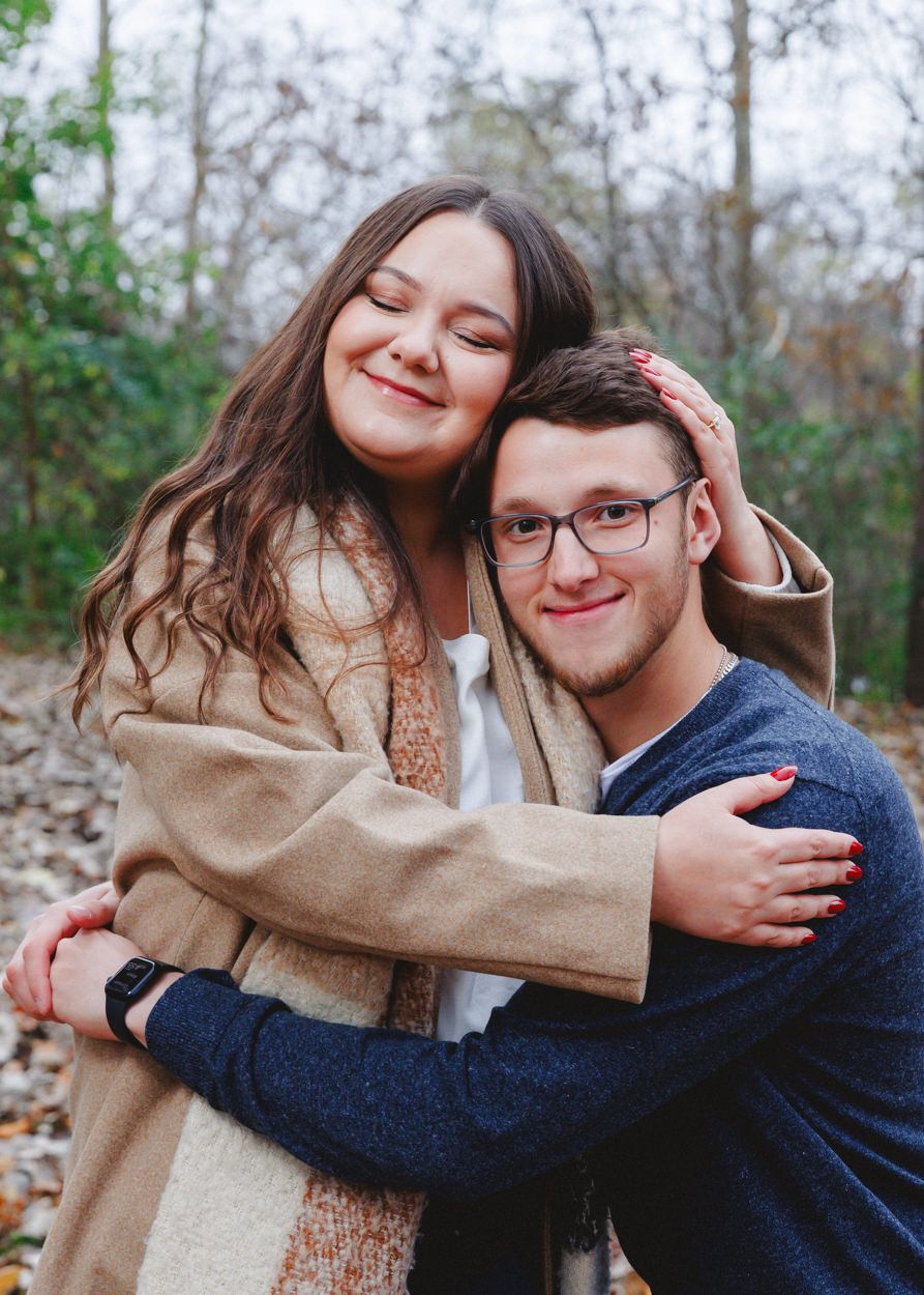 Romantic couple in warm sweaters share an intimate embrace while smiling in a forest setting.