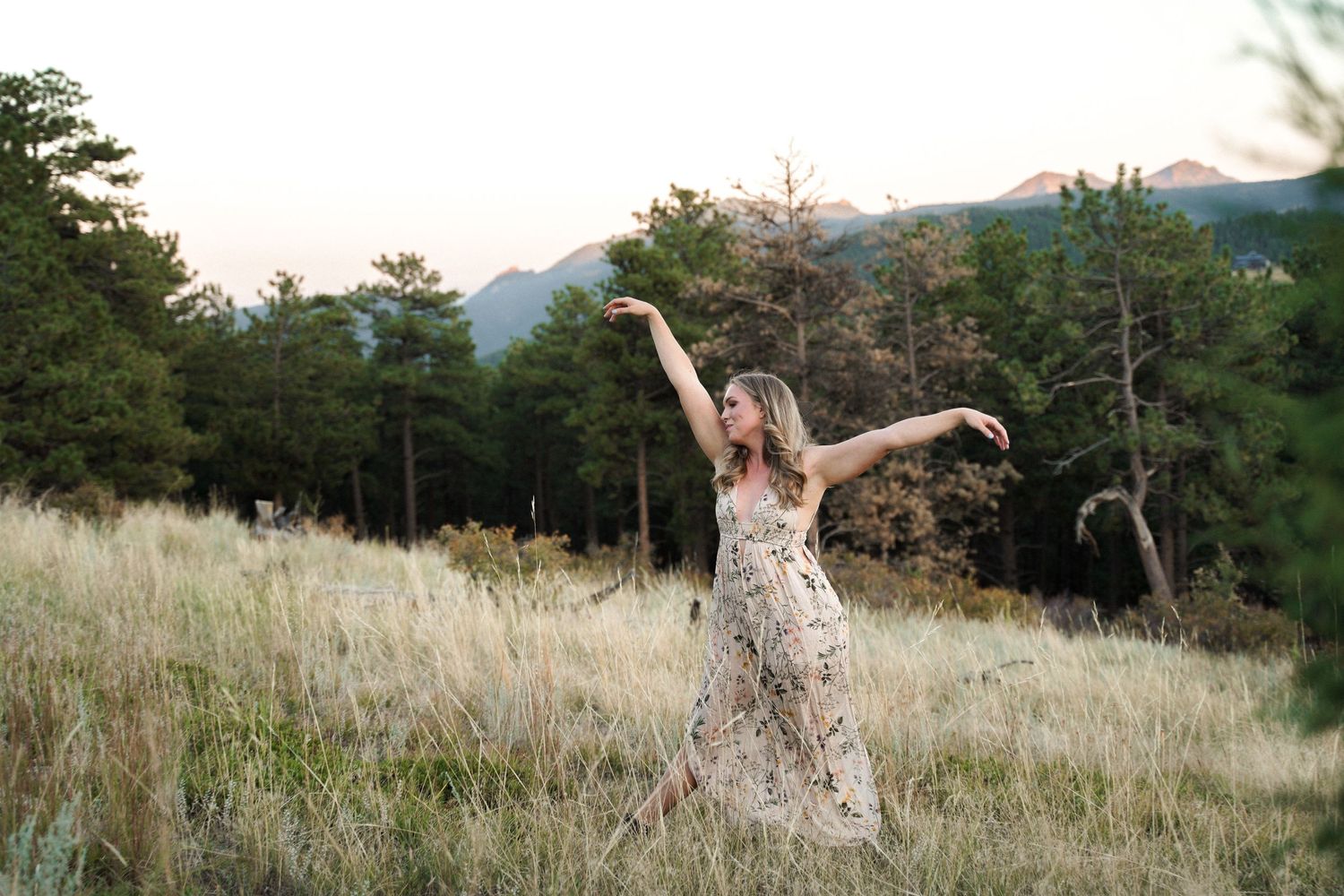 Woman in floral dress dancing with arms raised in mountain meadow at sunset.