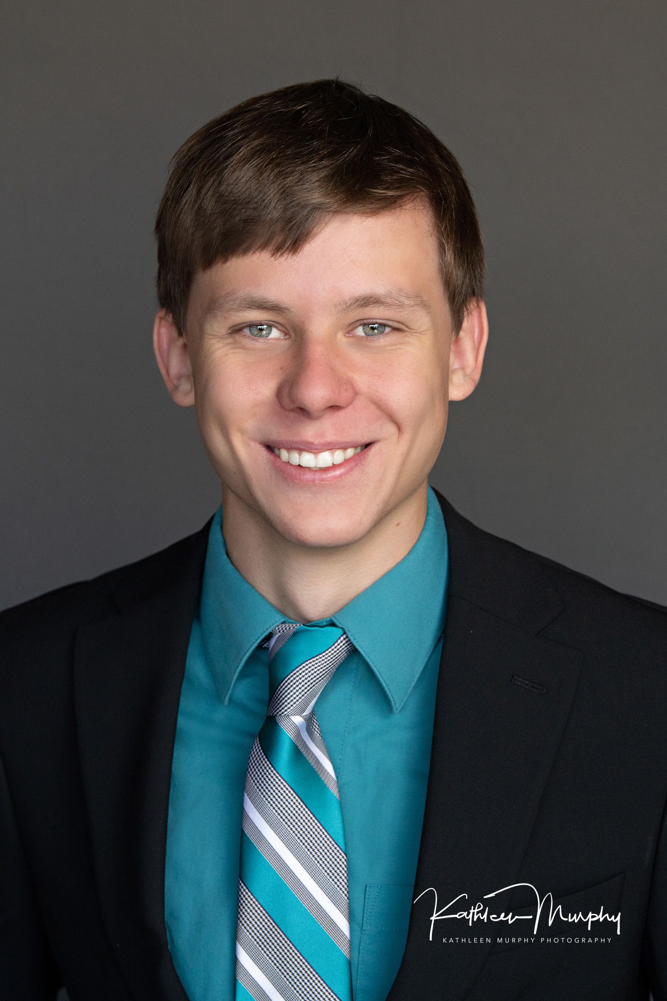 Professional headshot of person wearing black suit jacket with teal dress shirt and striped tie against grey background.