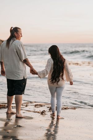 A couple walks hand-in-hand along a beach at sunset with waves crashing in the background.