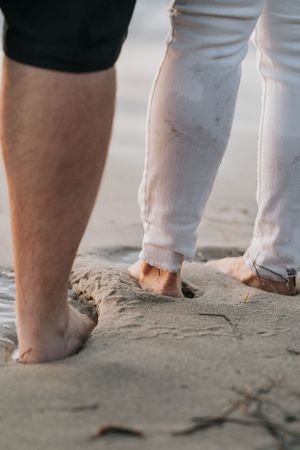 Close-up view of bare feet in sand on a beach with one person wearing white pants.