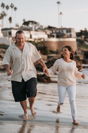 A couple runs joyfully together on a wet beach with coastal homes in the background.