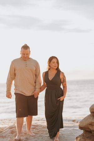 Couple walking barefoot along rocky coastal shoreline at sunset.