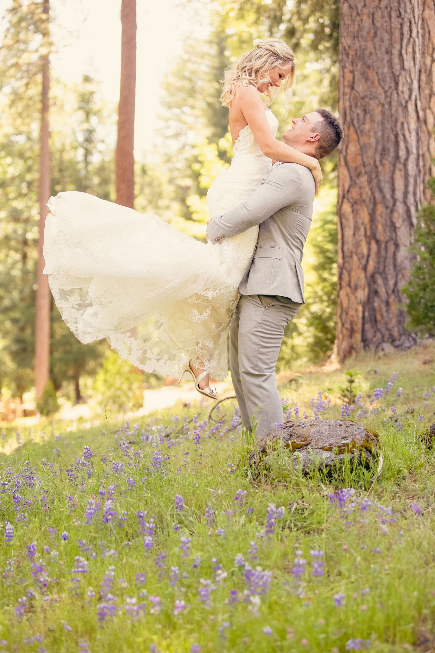 Groom lifting bride in white gown among purple wildflowers in a sunlit forest clearing.
