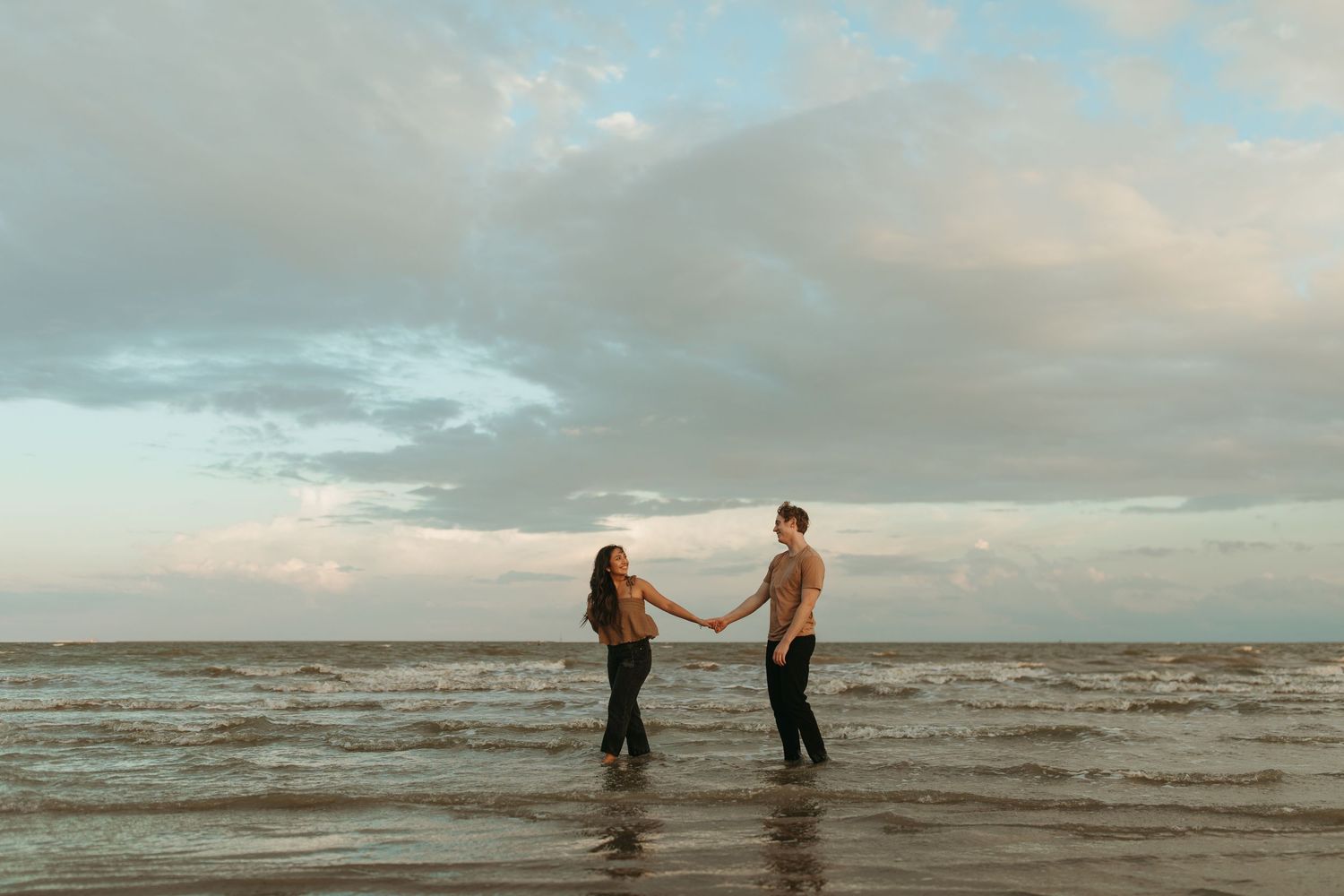 A couple standing together in shallow ocean water during sunset with cloudy skies.
