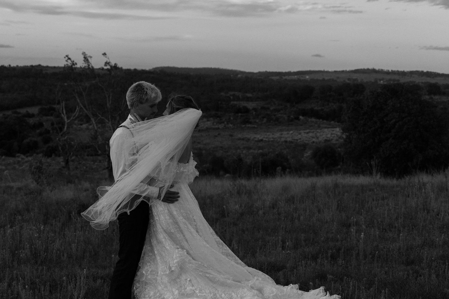 A romantic black and white photo of a couple with flowing wedding dress veil in a scenic hillside.