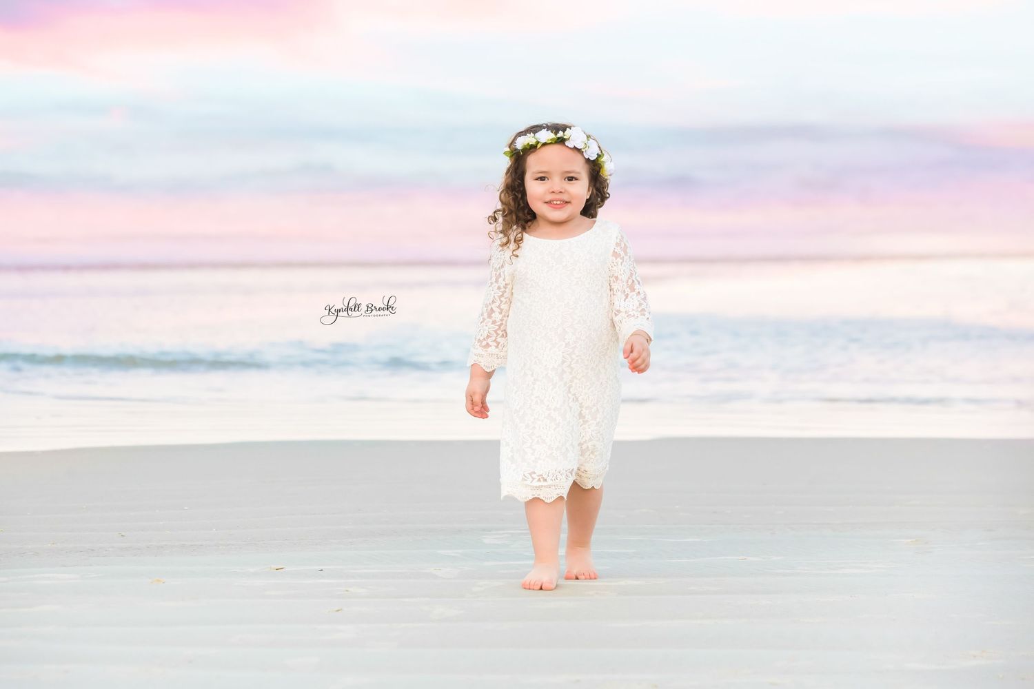 Small child in white lace dress walking on beach during sunset.