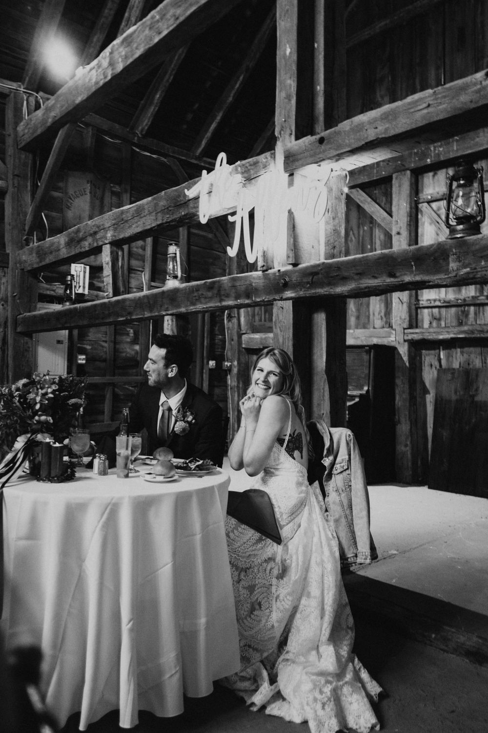 Black and white photo of wedding reception in rustic barn setting with exposed beams.