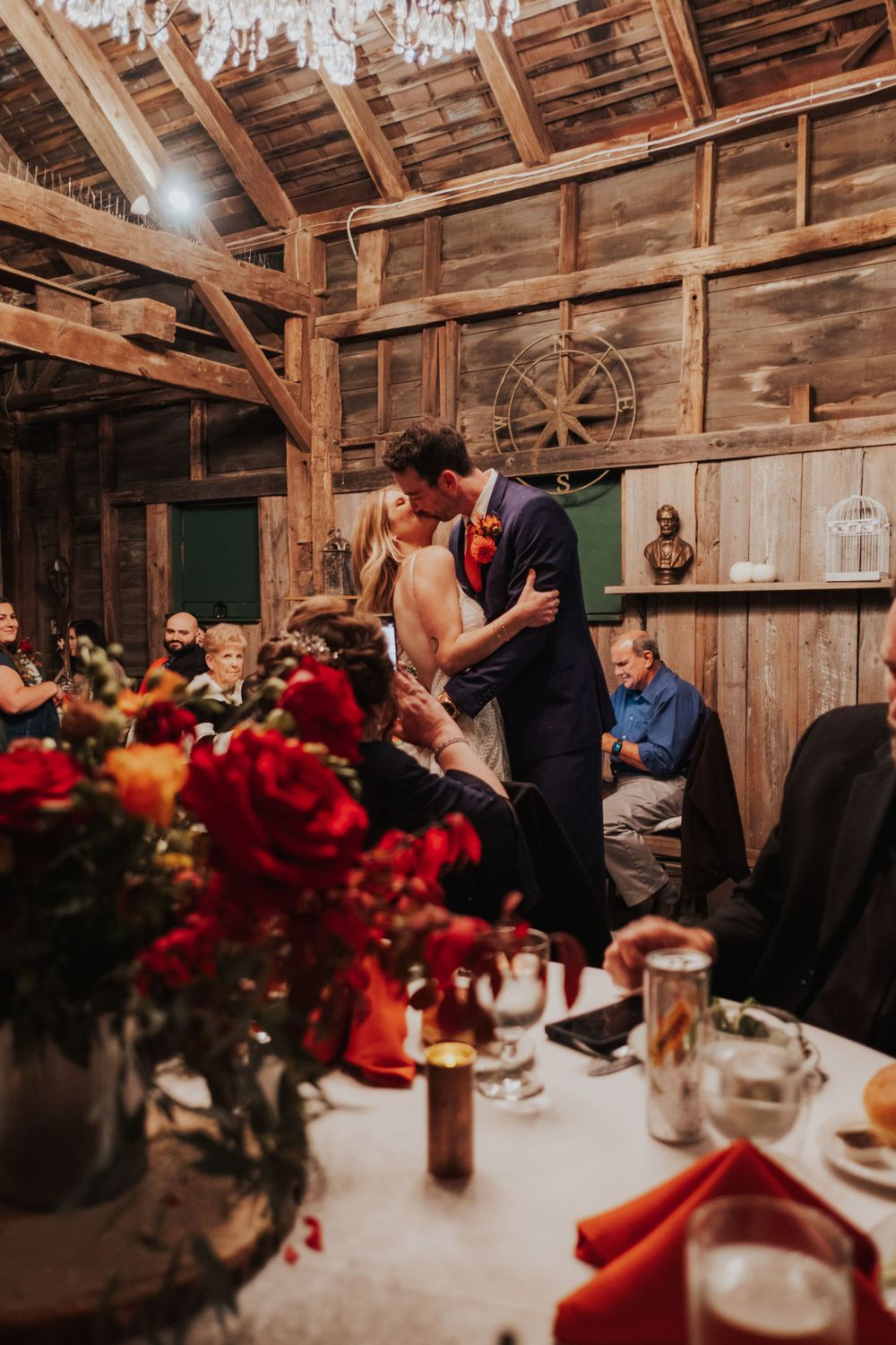 Intimate wedding reception moment in rustic barn with red floral centerpieces.