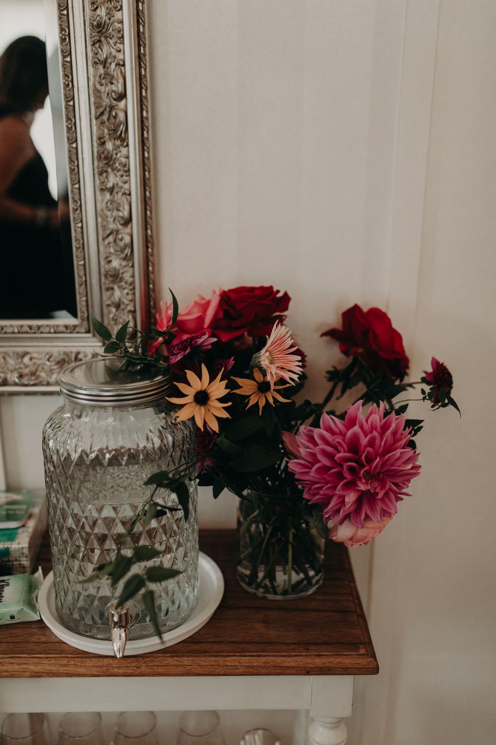Decorative glass jars and colorful dahlias displayed on wooden shelf with ornate mirror.