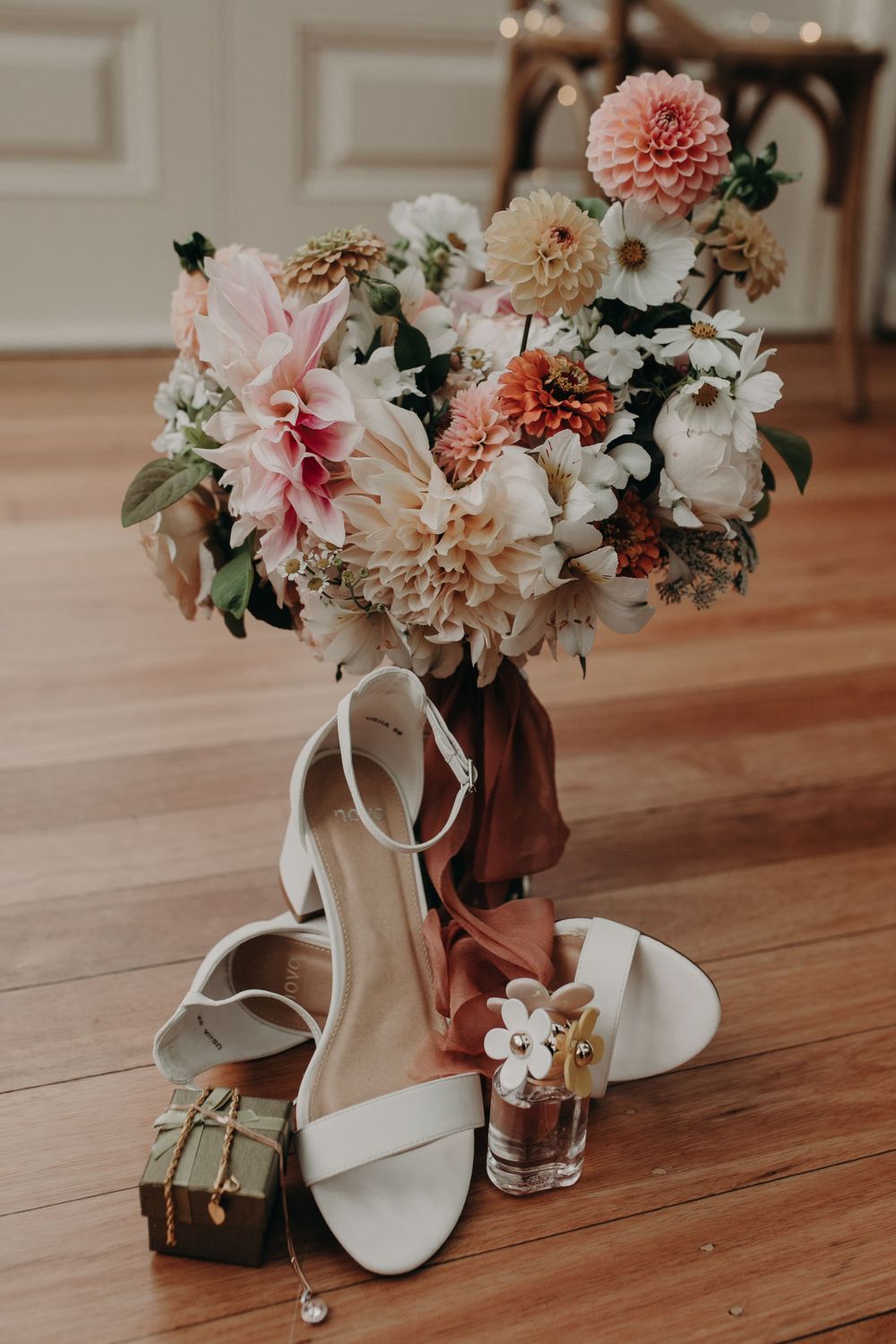 White bridal shoes next to pink and peach floral arrangements on hardwood floor.