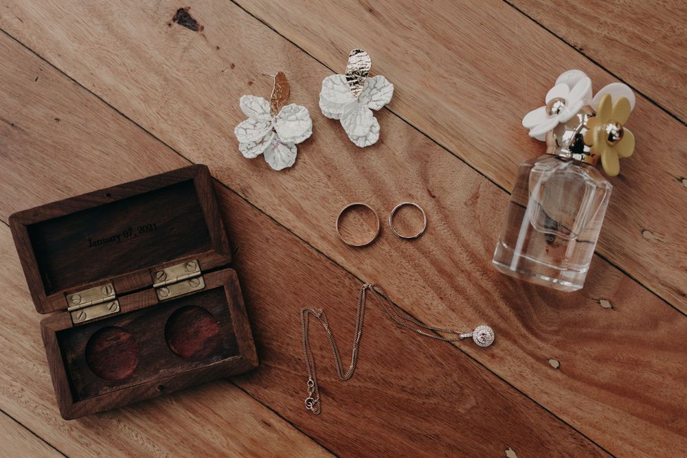 Wedding details including rings, white flowers, perfume bottle and wooden box on rustic wooden surface.